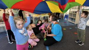 Children participating in a music and parachute play activity with an educator at Jacaranda Childcare Centre, encouraging creativity, movement, and social learning.